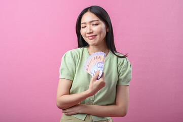 Fototapeta premium Asian woman in green top smiling confidently while holding a fan of purple Indonesian banknotes in one hand, standing against a solid pink background