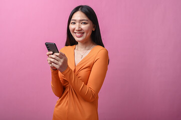 Naklejka premium Asian woman with long black hair in orange dress standing indoors against pink background, holding smartphone with both hands, smiling and looking forward with confident and relaxed posture.