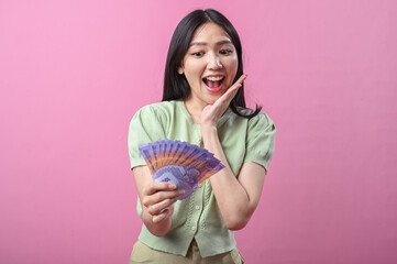 Asian woman in green top looks amazed while holding a fan of Indonesian rupiah banknotes in front...