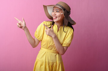 Asian woman in a yellow dress and sun hat smiling while pointing to the side with both hands,...