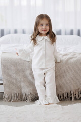 Child in white pajamas standing on bed beside cozy blanket in bright room