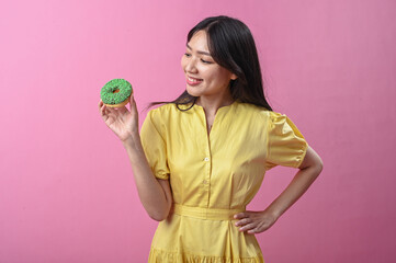Asian woman in a yellow dress smiling while holding a green-sprinkled donut in front of a pink...