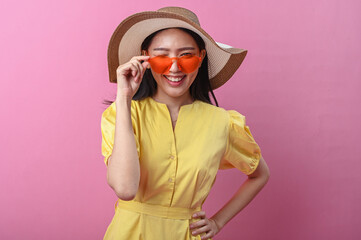 Asian woman in a yellow dress and wide-brimmed hat smiling playfully while holding orange sunglasses, posing against a solid pink background with a cheerful expression