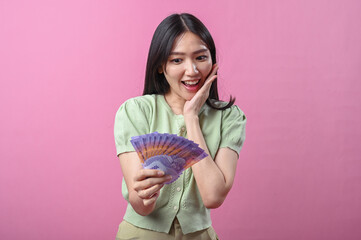 Asian woman in green top looks amazed while holding a fan of Indonesian rupiah banknotes in front...