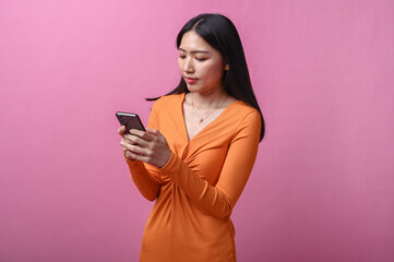 Asian woman with long black hair wearing a fitted orange dress stands indoors against pink...