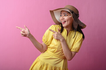 Asian woman in a yellow dress and sun hat smiling while pointing to the side with both hands,...