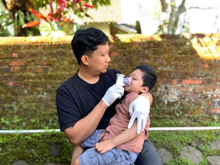 A father wearing medical gloves holds his sick child and gives him nebulizer therapy in an outdoor...