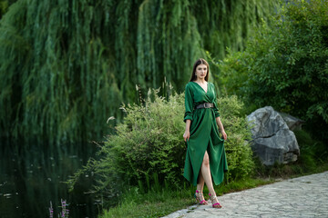 Elegant woman in a green dress walking by a lake on a stone path, surrounded by lush greenery and...