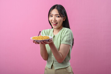 Asian woman in light green top smiling while presenting a plate of cooked instant noodles with a...