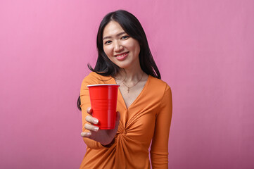 Asian woman with long black hair in orange dress smiling while extending a red plastic cup toward...