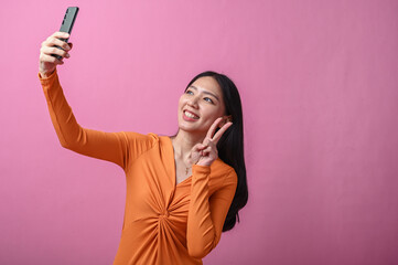 Asian woman with long black hair wearing an orange dress posing for selfie with smartphone against...