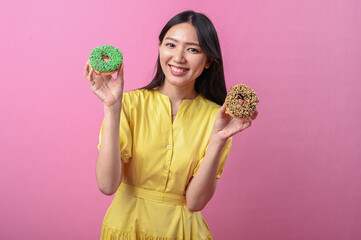 Asian woman in a yellow dress smiling brightly while holding two decorated donuts, one green and...