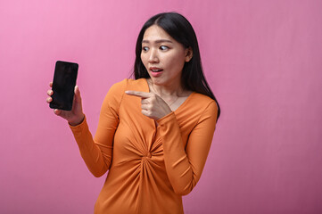 Asian woman with long black hair in orange dress smiling and pointing at smartphone screen with...