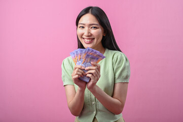 Asian woman in green top smiling happily while holding a fan of purple banknotes in both hands,...