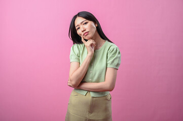 Asian woman thinking with hand on chin, looking up thoughtfully while standing against a plain pink...