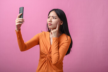 Asian woman with long black hair wearing orange dress holding smartphone and looking confused with...