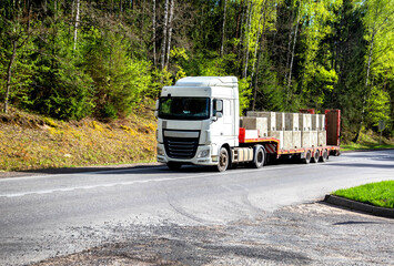 Modern truck with a lowboy semitrailer transports concrete blocks along a country road against the...