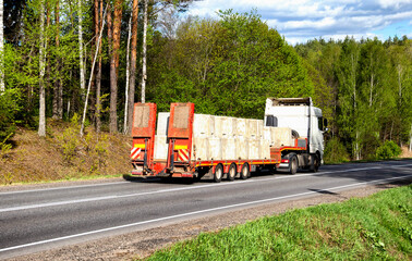 Modern truck with a lowboy semitrailer transports concrete blocks along a country road against the...