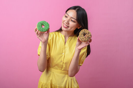 Asian woman in a yellow dress smiling brightly while holding two decorated donuts, one green and one chocolate with nuts, posing cheerfully against a pink background - Powered by Adobe