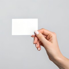 A minimalist photograph featuring a single, fair-skinned hand holding a blank white card against a plain light gray background. The hand is manicured with light-colored nail polish.