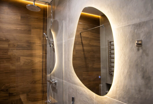 Modern mirror with yellow LED backlight in the bathroom against the background of a chrome shower and brown wood tiles. Copy space for text