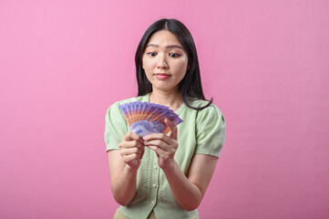 Asian woman looking surprised and excited while holding a fan of Indonesian rupiah banknotes,...