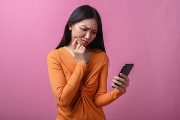 Asian woman with long black hair wearing orange dress looking confused while holding smartphone in...