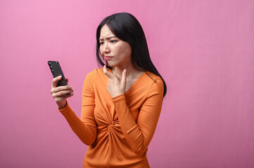 Asian woman with long black hair wearing orange dress holding smartphone and looking confused with...