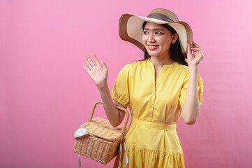 Asian woman in a yellow dress smiling warmly while waving and holding a wicker picnic basket. She...