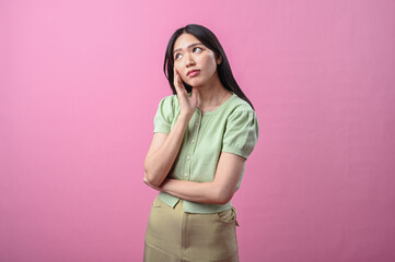 Asian woman thinking with hand on chin, looking up thoughtfully while standing against a plain pink...