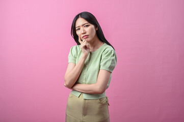 Asian woman thinking with hand on chin, looking up thoughtfully while standing against a plain pink...