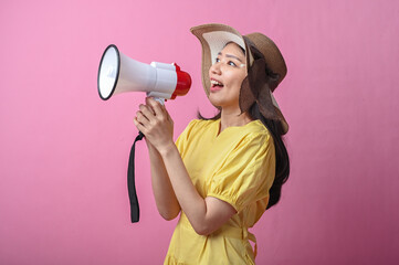 Asian woman in yellow dress and sun hat cheerfully shouting into a megaphone, standing against a...