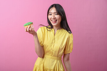 Asian woman in a yellow dress smiling while looking at a green-sprinkled donut in her hand, posing...