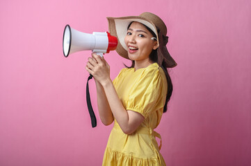 Asian woman in yellow dress and sun hat cheerfully shouting into a megaphone, standing against a...