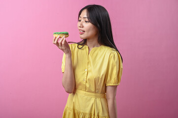 Asian woman in a yellow dress about to bite a green-sprinkled donut while looking at the camera,...