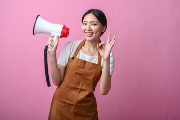 Asian woman in brown apron smiling while holding a megaphone and making an OK hand gesture,...