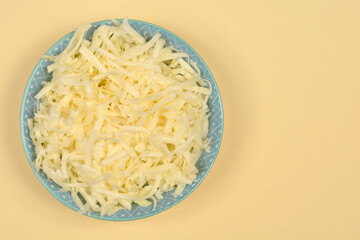 An overhead shot displays a light blue patterned bowl filled with freshly shredded cheese, positioned on the left side of a soft beige background with ample copy space