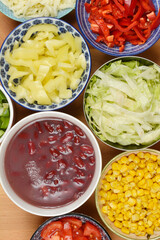 An overhead flat lay showcases a vibrant assortment of fresh, chopped salad ingredients, including lettuce, bell peppers, corn, and beans, presented in various bowls on a wooden surface