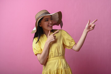 Asian woman in a yellow dress and sun hat smiling while pointing to the side with both hands,...