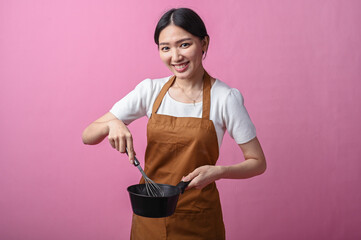 Asian woman in brown apron stirring ingredients in a black saucepan with a whisk, smiling in front...
