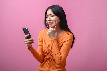 Asian woman with long black hair wearing orange dress holding smartphone and looking confused with...