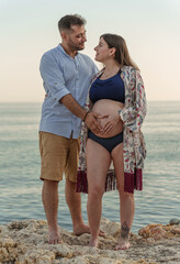 A young heterosexual couple consisting of a man and a pregnant woman posing on the edge of a cliff, with the sea in the background, as they gaze lovingly at each other and hold hands on her belly.