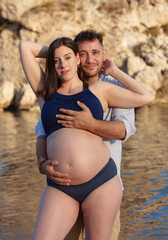Beautyful pregnant woman posing in a bikini, with her husband hugging her from behind, on the shore at sunset.