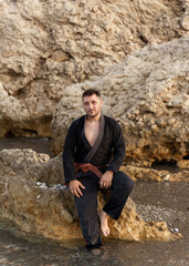 adult male martial arts practitioner wearing a black kimono, sitting on a large rock on the shore of the beach, with his feet in the water and a large rock behind him.