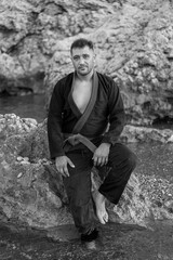 Black and white portrait of an adult male karateka wearing a kimono, sitting on a large rock on the shore of the beach, with his feet in the water and a large rock behind him.
