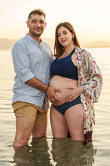 Attractive young couple in love, posing in the waters of the beach shore, both holding the pregnant woman's belly, at sunset
