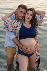 An attractive young couple expecting a child pose happily and smilingly embracing each other in the calm waters of the sea, near the shore of the beach, with the father's hands on the pregnant woman's