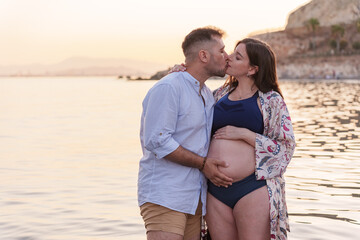 An attractive young couple kissing in the calm waters of the sea, near the shore of the beach, both holding the pregnant woman's belly with one hand, at sunset.