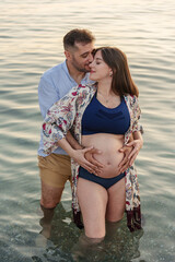 A young couple in love, expecting a child, posing happy and smiling as they embrace and kiss in the calm waters of the sea, near the shore of the beach, with their hands on the pregnant woman's belly.