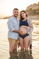 A young couple expecting a child posing happily and smiling in the calm waters of the sea, near the shore of the beach, both holding the pregnant woman's belly with one hand, at sunset.